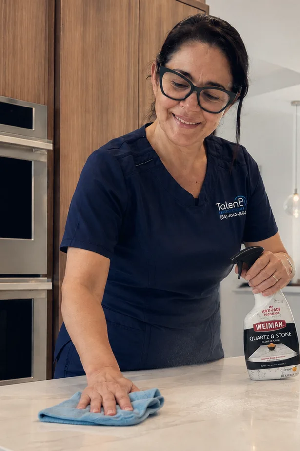 A Talent Cleaning Services professional wiping down a kitchen counter beside built-in ovens, with the living room visible behind her.