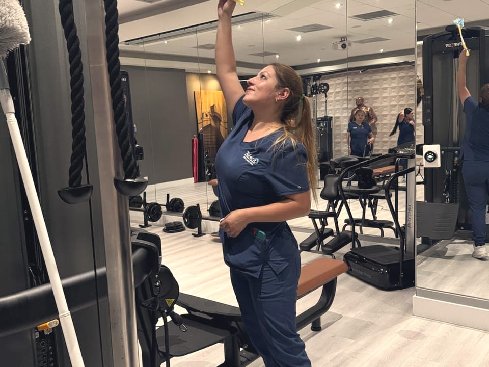 A Talent Cleaning Services professional dusting the top of a gym machine with an extension pole, in front of the mirrored wall.