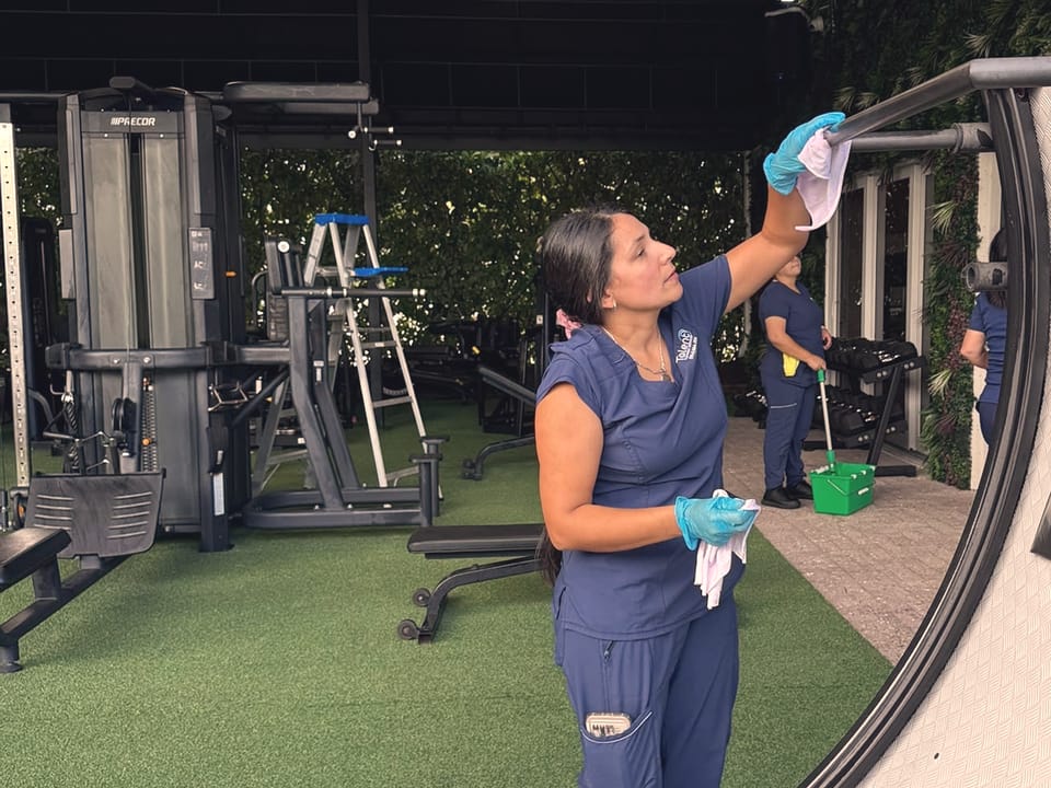 A Talent Cleaning Services professional in gloves wiping the metal frame of an open-air gym rig with a cloth, with a colleague mopping the walkway behind her.