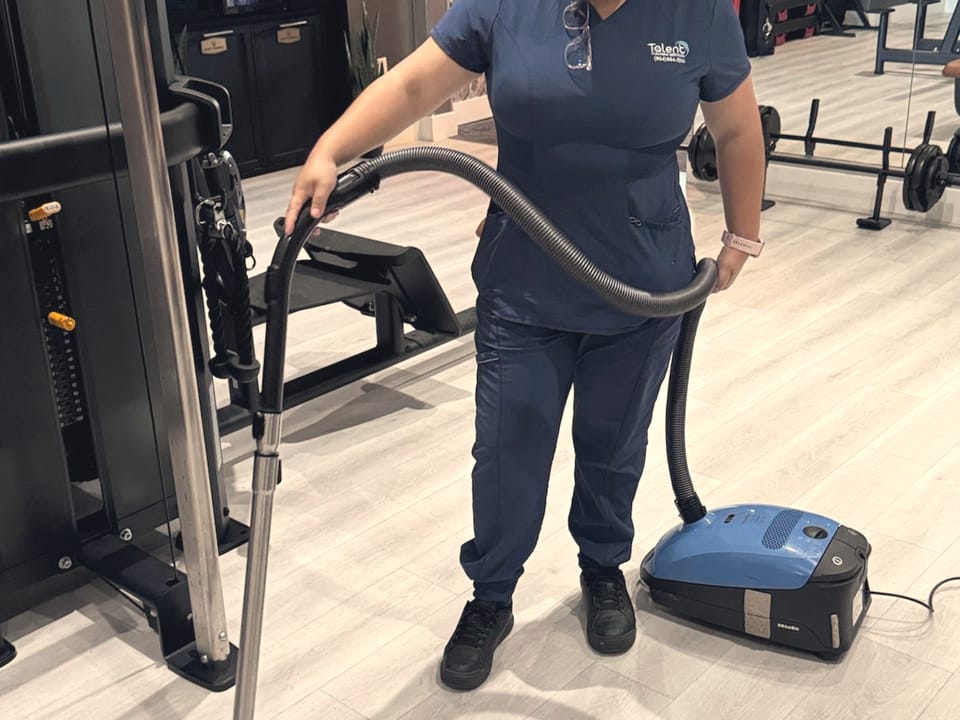 A Talent Cleaning Services professional vacuuming the floor of a gym between the weight machines, with the canister vacuum beside her.