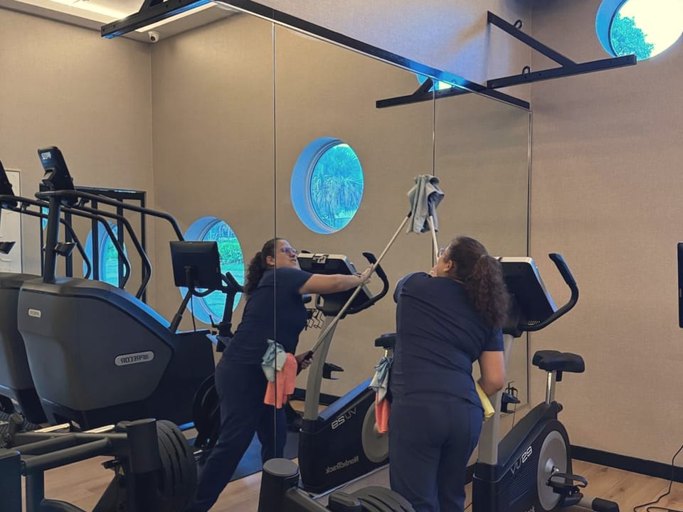 A Talent Cleaning Services professional dusting a high surface with a cloth on a pole, her reflection in the mirrored wall beside the exercise bikes.