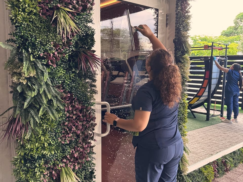 A Talent Cleaning Services professional pulling a squeegee down a glass door framed by a living plant wall, with a colleague cleaning another surface outside.
