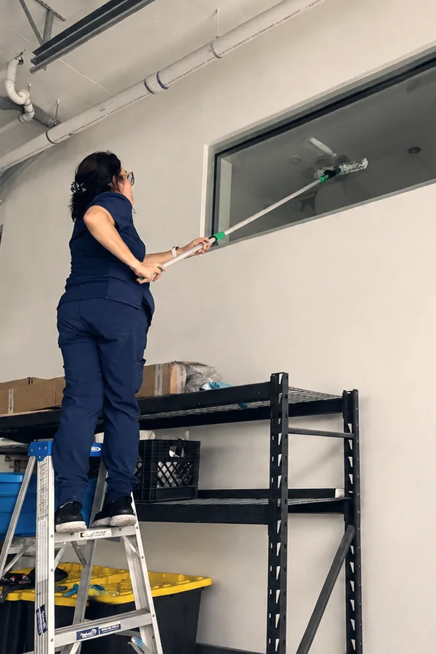 A Talent Cleaning Services professional on a step ladder cleaning a high window with an extension pole, in a warehouse with cleaning equipment on the shelving below.